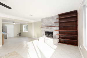Unfurnished living room featuring ceiling fan, light tile patterned flooring, a stone fireplace, and ornamental molding