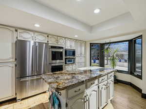 Kitchen with a tray ceiling, appliances with stainless steel finishes, a center island, light wood-style flooring, and dark stone counters
