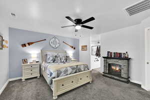 Bedroom featuring a stone fireplace, a ceiling fan, dark carpet, and ensuite bath