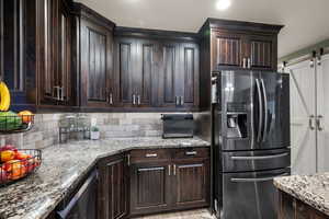 Kitchen with stainless steel fridge, dark brown cabinetry, light stone countertops, and backsplash