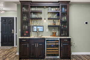 Indoor wet bar with dark brown cabinets, wine cooler, light stone countertops, and open shelves
