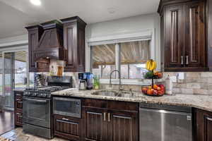 Kitchen with dark brown cabinets, appliances with stainless steel finishes, light stone counters, custom range hood, and decorative backsplash