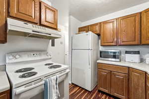Kitchen with white appliances, brown cabinetry, light countertops, under cabinet range hood, and dark wood-style flooring