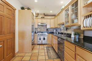 Kitchen featuring premium appliances, dark stone counters, glass insert cabinets, under cabinet range hood, and light brown cabinetry