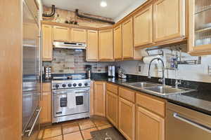 Kitchen with high quality appliances, under cabinet range hood, dark stone counters, light brown cabinets, and glass insert cabinets