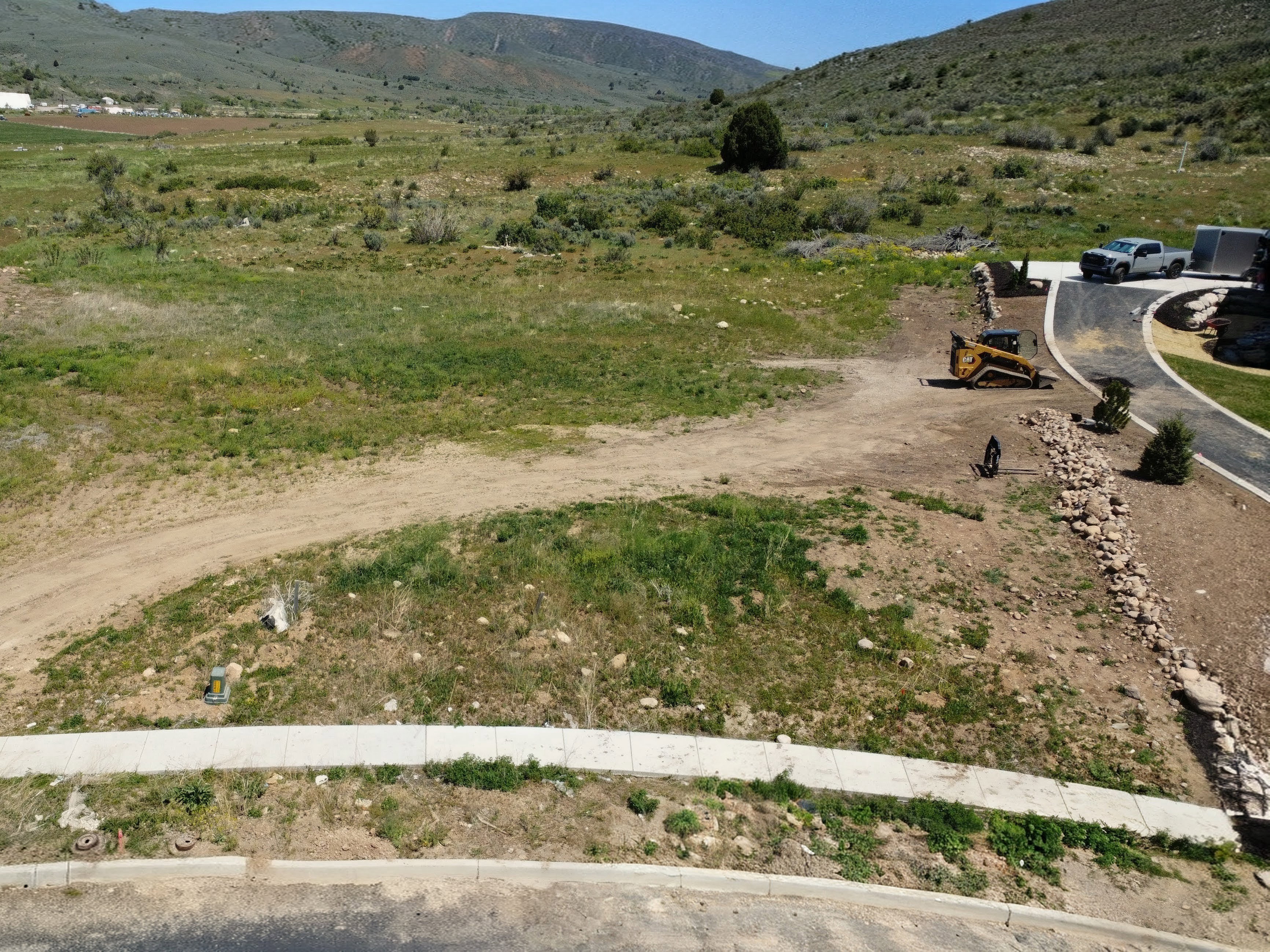Expansive undeveloped lot featuring a paved driveway, a retaining wall composed of natural stones, a concrete sidewalk, and surrounding natural terrain with distant hills