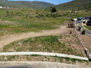 Expansive undeveloped lot featuring a paved driveway, a retaining wall composed of natural stones, a concrete sidewalk, and surrounding natural terrain with distant hills