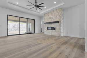 Unfurnished living room featuring light wood-type flooring, a stone fireplace, a ceiling fan, a raised ceiling, and recessed lighting