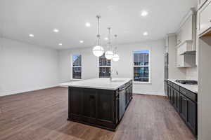 Kitchen with an island with sink, recessed lighting, decorative light fixtures, dark wood-style flooring, and light stone counters