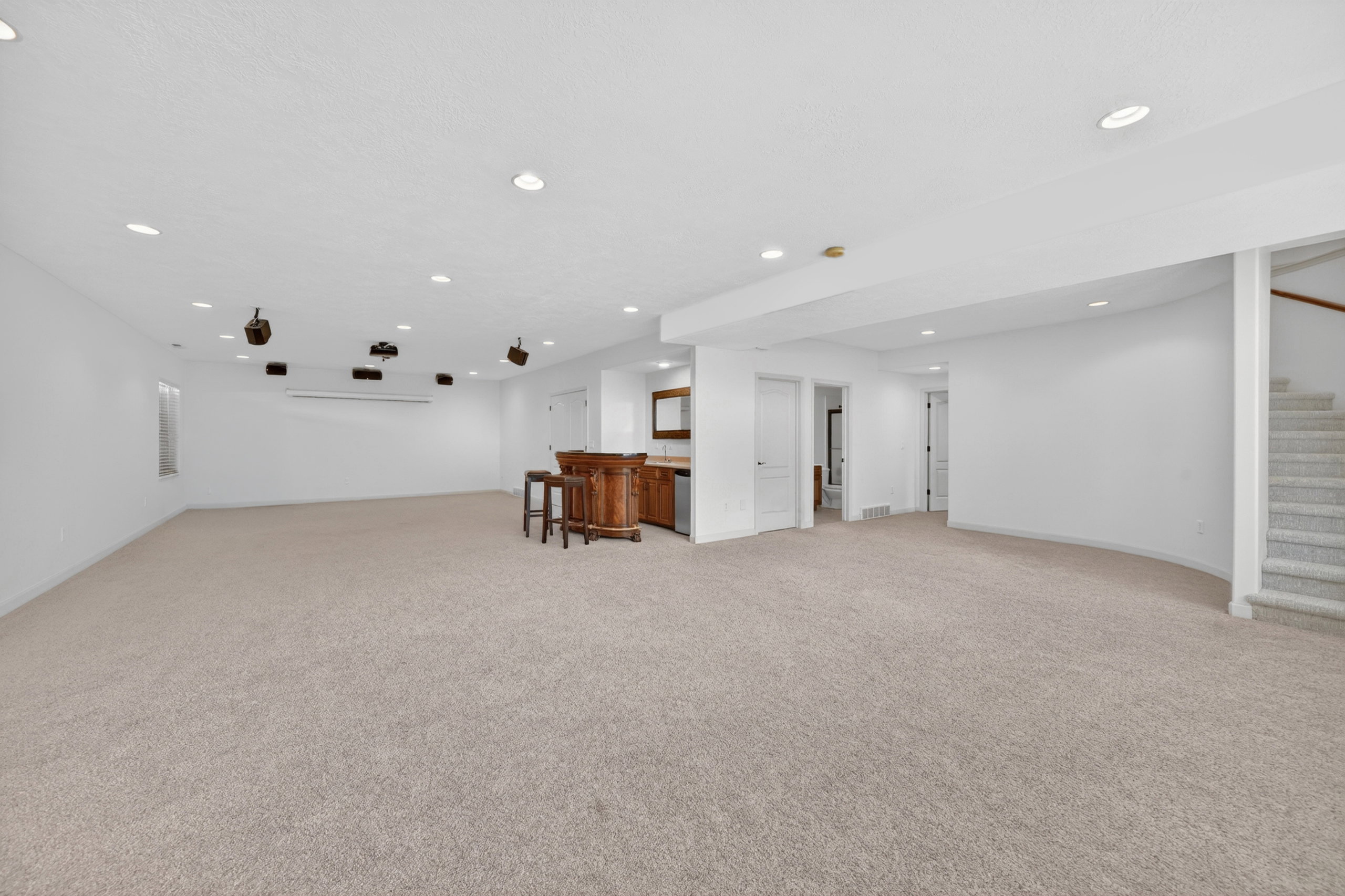 Spacious carpeted room featuring recessed lighting, an integrated home theater system with screen and speakers, a built-in wet bar with wooden cabinetry, and a carpeted staircase with a wood handrail
