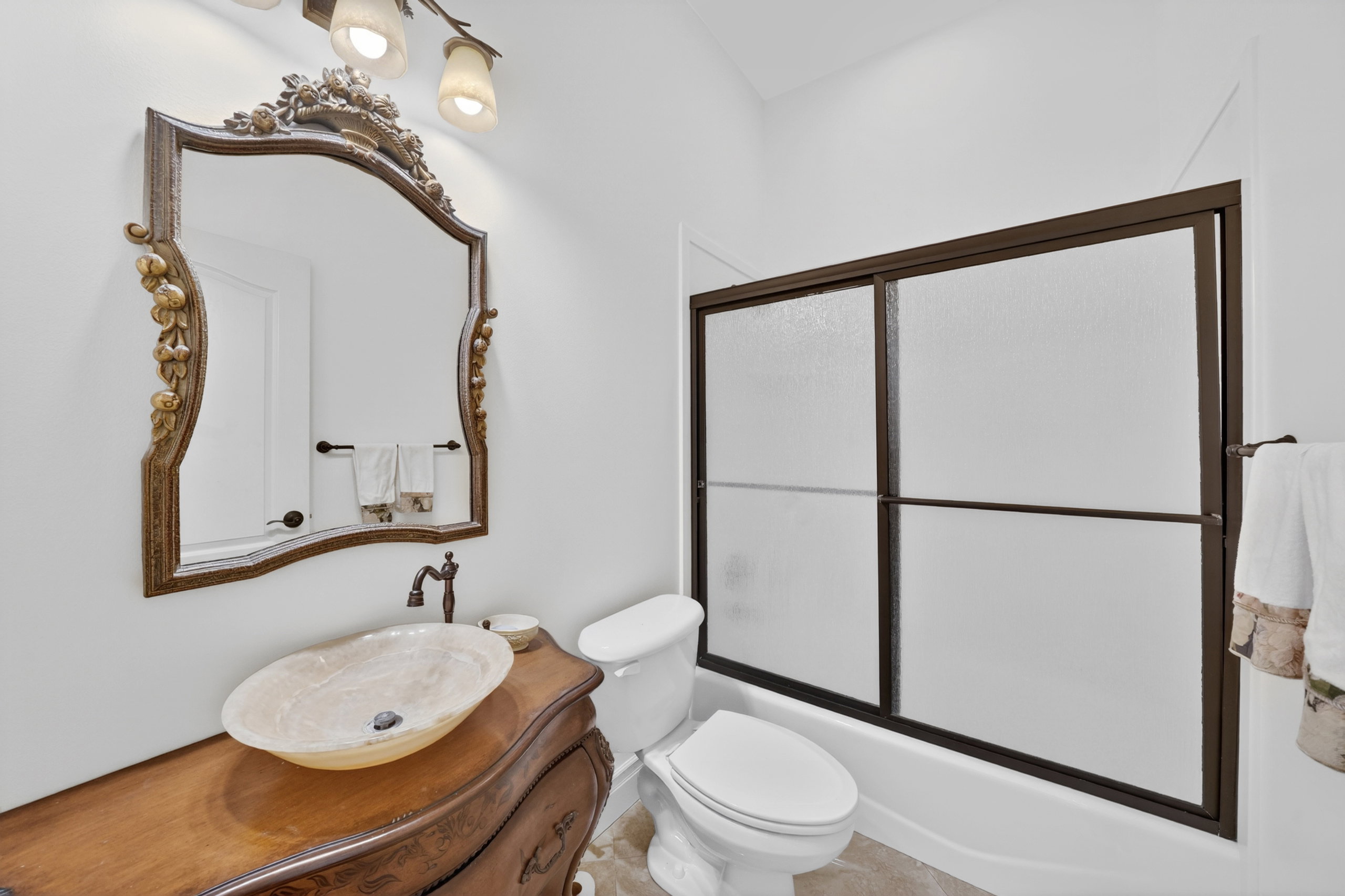 Bathroom featuring a decorative carved mirror, an oval vessel sink, a wood-finish vanity, a toilet, and a tub/shower combination with sliding frosted glass doors