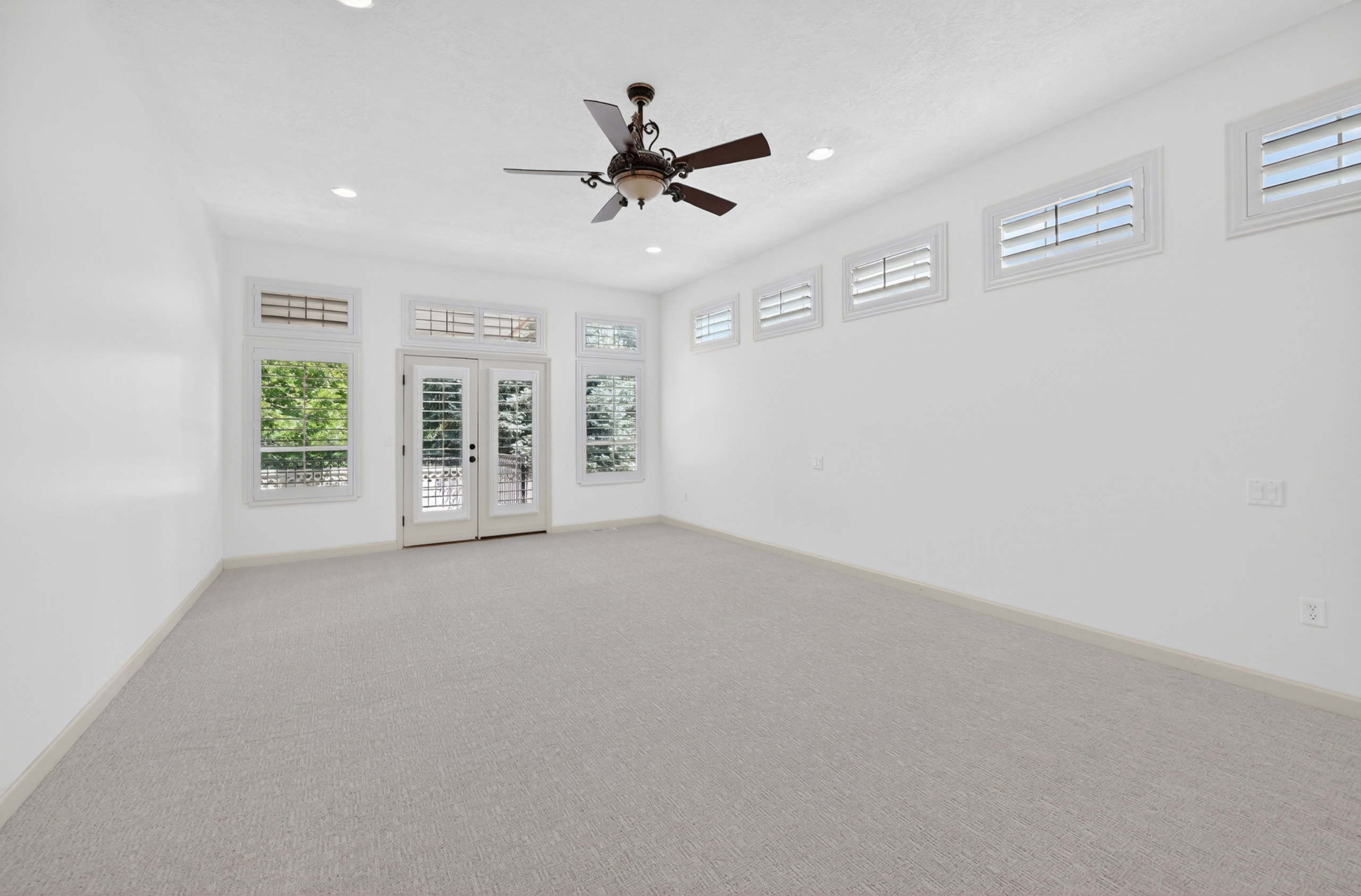 Spacious room featuring light-toned carpet flooring, a ceiling fan, recessed lighting, and multiple windows with white plantation shutters