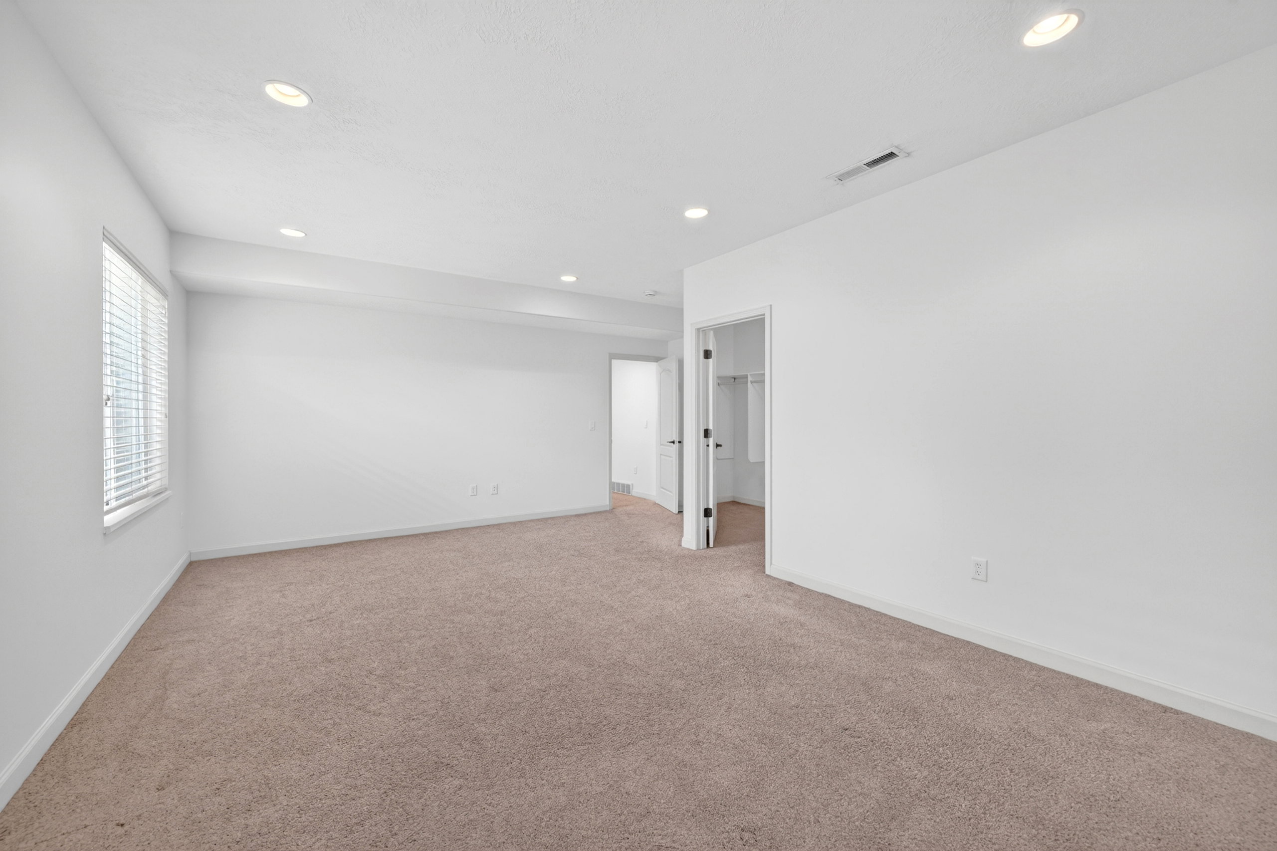 Spacious room featuring neutral carpeting, white walls, recessed ceiling lighting, a window with blinds, and a doorway leading to an additional space with shelving