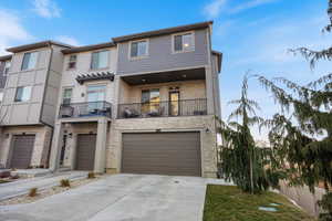 View of front facade featuring a balcony, stone siding, driveway, and an attached garage