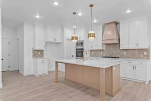 Kitchen with decorative backsplash, custom range hood, appliances with stainless steel finishes, light stone countertops, and white cabinetry