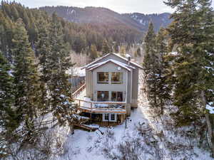 Snow covered back of property featuring a deck with mountain view and a wooded view