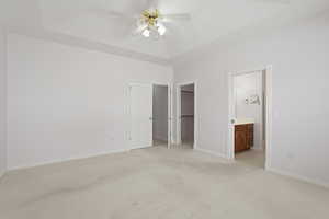 Unfurnished bedroom featuring light colored carpet, a walk in closet, a ceiling fan, ensuite bath, and a tray ceiling
