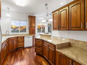 Kitchen with light stone countertops, dark wood-style flooring, hanging light fixtures, white dishwasher, and brown cabinets