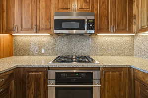 Kitchen with stainless steel appliances, light stone countertops, brown cabinets, and tasteful backsplash