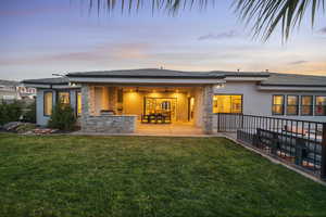 Back of house at dusk featuring a ceiling fan, stucco siding, exterior kitchen, a yard, and stone siding