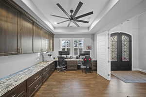 Home office with a raised ceiling, french doors, ceiling fan, built in desk, and dark wood-style flooring