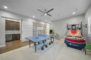Game room featuring a barn door, ceiling fan, beverage cooler, light marble finish floors, and bar with sink