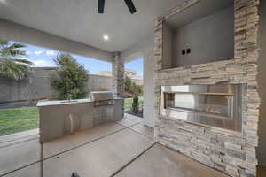 Fenced backyard with an outdoor kitchen, a patio area, a ceiling fan, and an outdoor stone fireplace