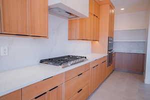Kitchen with light stone counters, exhaust hood, modern cabinets, light brown cabinetry, and appliances with stainless steel finishes