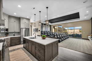 Kitchen with a raised ceiling, stainless steel appliances, hanging light fixtures, a center island with sink, and light stone counters