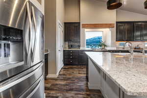 Kitchen featuring stainless steel appliances, light stone counters, dark wood-style floors, vaulted ceiling, and dark brown cabinets