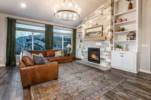 Living room with built in shelves, dark wood-style flooring, a stone fireplace, lofted ceiling, and a chandelier
