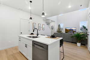 Kitchen with hanging light fixtures, stainless steel dishwasher, white cabinetry, a center island with sink, and light wood-type flooring