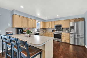 Kitchen with appliances with stainless steel finishes, light brown cabinets, light countertops, a kitchen breakfast bar, and dark wood finished floors