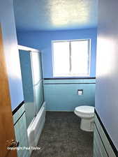 Full bathroom with tile walls, a textured ceiling, a wainscoted wall, and bath / shower combo with glass door