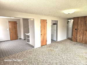 Unfurnished bedroom with a textured ceiling, carpet flooring, freestanding refrigerator, a closet, and wooden walls