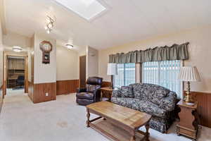 Living room with wooden walls, wainscoting, a desk, light colored carpet, and a skylight