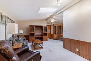 Carpeted living room featuring wooden walls, wainscoting, a skylight, and a ceiling fan