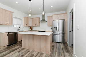 Kitchen featuring light brown cabinets, appliances with stainless steel finishes, wall chimney range hood, hanging light fixtures, and a kitchen island