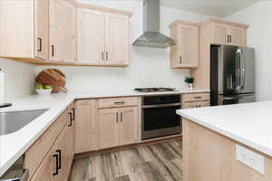 Kitchen with light brown cabinetry, appliances with stainless steel finishes, wall chimney range hood, light wood finished floors, and light stone countertops