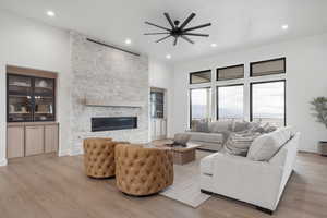 Living area with light wood-style flooring, a stone fireplace, a ceiling fan, a high ceiling, and recessed lighting