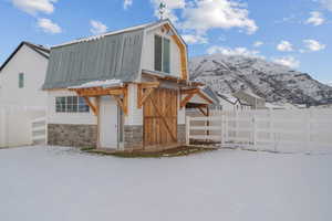 Snow covered property featuring a gambrel roof, a fenced backyard, stone siding, an outdoor structure, and a metal roof