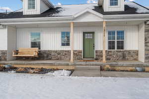 Snow covered property entrance with covered porch, roof with shingles, stone siding, and board and batten siding