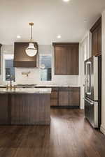 Kitchen featuring high end refrigerator, custom exhaust hood, pendant lighting, dark brown cabinets, and dark wood-style flooring