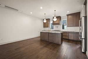 Kitchen with a center island with sink, high end fridge, hanging light fixtures, premium range hood, and dark brown cabinets