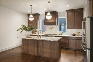 Kitchen featuring high end fridge, an island with sink, dark wood-style flooring, custom exhaust hood, and decorative light fixtures