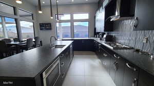 Kitchen with dark cabinets, a center island with sink, wall chimney exhaust hood, decorative light fixtures, and stainless steel appliances