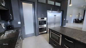 Kitchen featuring dark cabinetry, dark stone counters, built in appliances, light tile patterned flooring, and glass insert cabinets