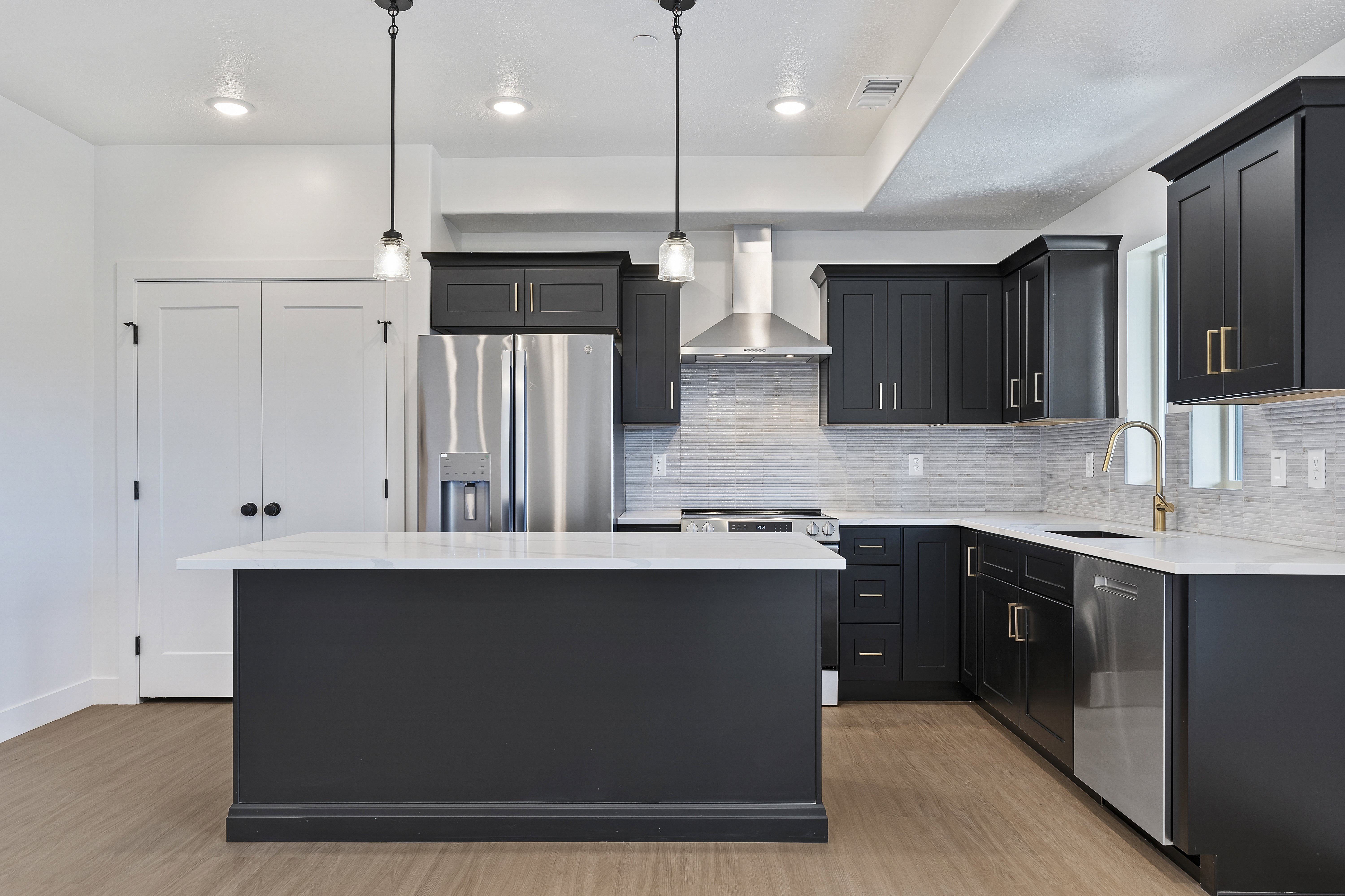 Kitchen with dark cabinets, appliances with stainless steel finishes, light stone counters, decorative light fixtures, and wall chimney range hood