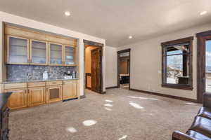 Indoor wet bar featuring glass insert cabinets, tasteful backsplash, light stone countertops, light colored carpet, and recessed lighting