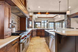 Kitchen featuring stainless steel appliances, decorative light fixtures, a tray ceiling, a center island with sink, and custom exhaust hood